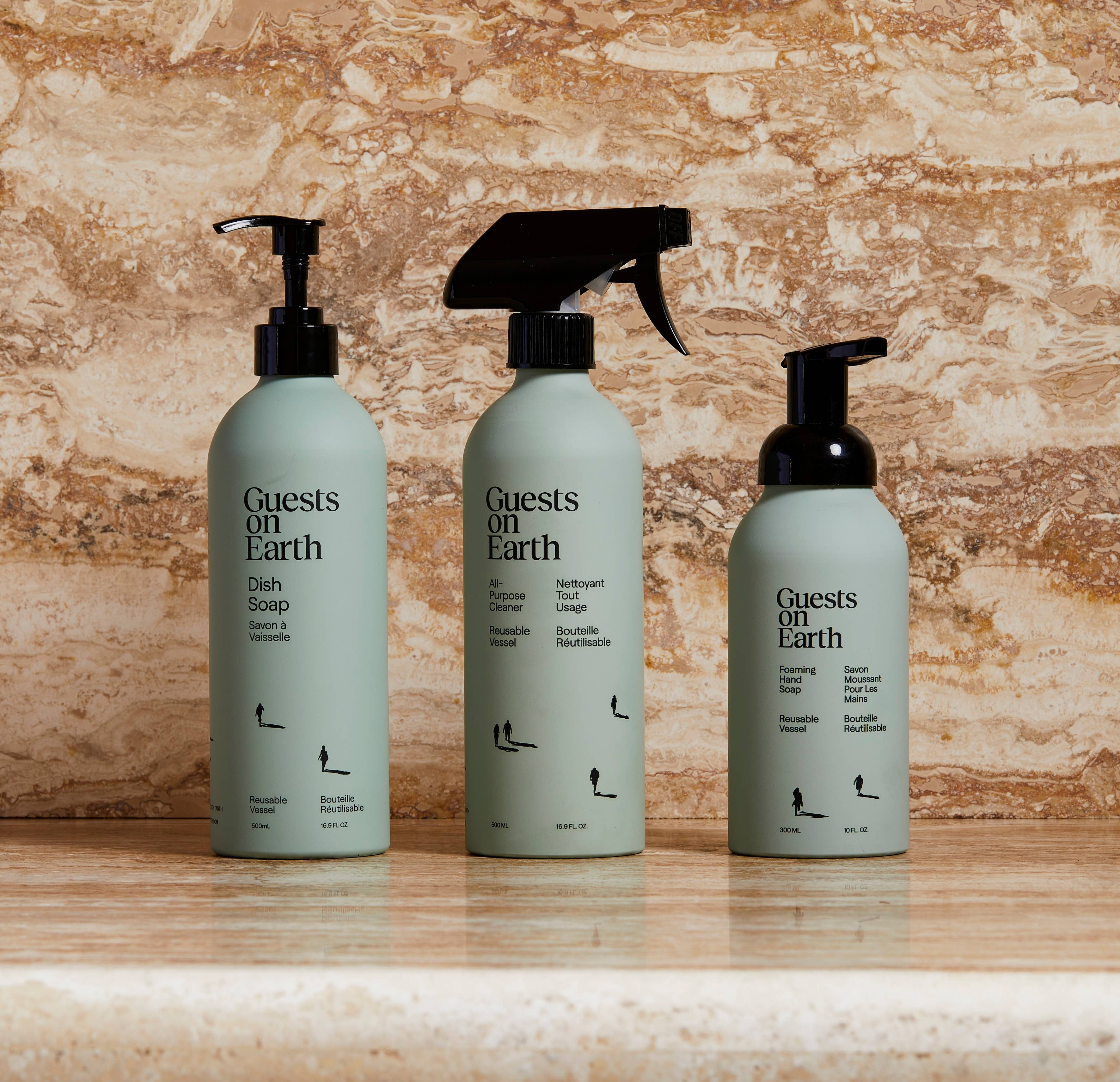 Three bottles of 'Guests on Earth' cleaning products on a wooden surface with a stone wall background.