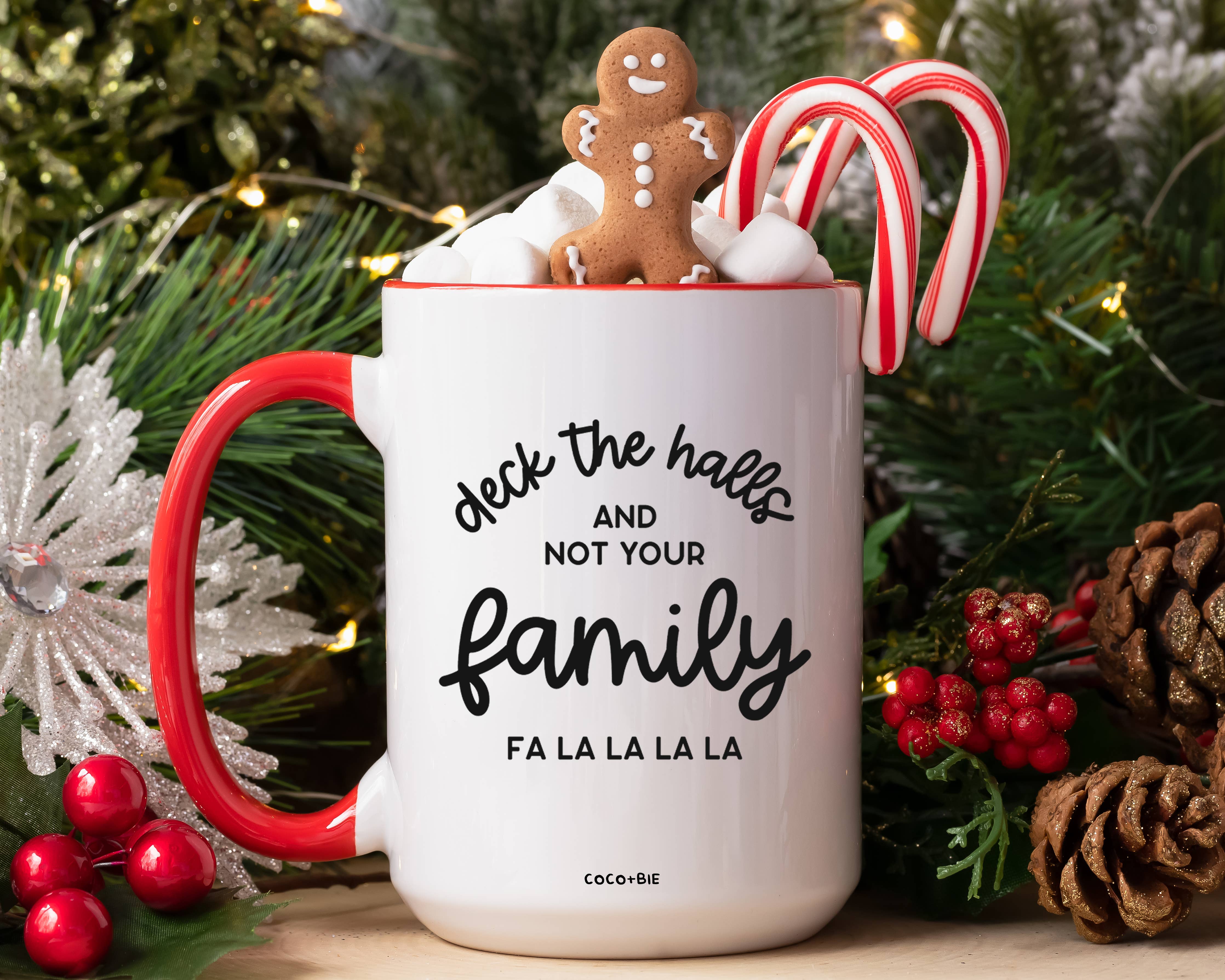 A white ceramic holiday mug with a red handle and red rim, printed with the phrase 'deck the halls AND NOT YOUR family FA LA LA LA LA'. The mug is styled with a gingerbread man, marshmallows, and candy canes, surrounded by Christmas greenery, pinecones, and red berries.