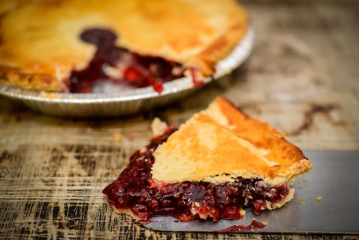 A slice of berry pie with a golden flaky crust and dark red berry filling, shown on a metal spatula over a rustic wooden surface, with the whole pie blurred in the background.