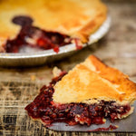 A slice of berry pie with a golden flaky crust and dark red berry filling, shown on a metal spatula over a rustic wooden surface, with the whole pie blurred in the background.