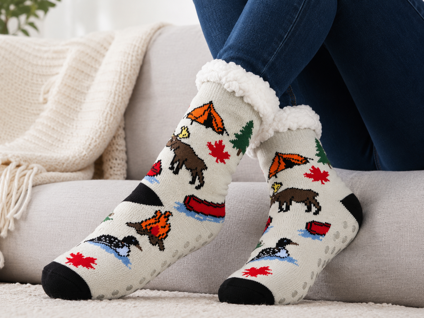 A person wearing cream knit socks with a colorful outdoor camping pattern, sitting on a light gray couch with a cream knit blanket.