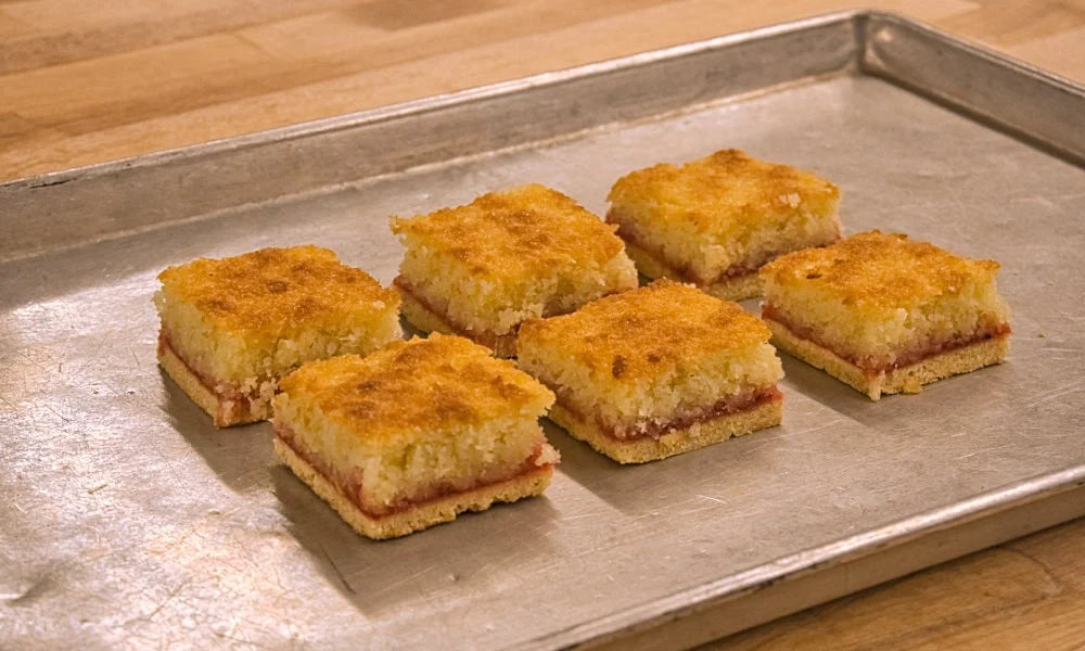 Six golden-brown baked bars with a crumbly topping and a red fruit filling, arranged on a metal baking sheet over a wooden surface.